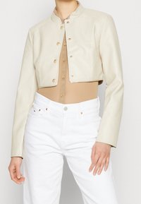 Veste en cuir beige raccourcie avec une fermeture à boutons-pression à l'avant, dotée d'un col rond, de manches longues et d'une texture lisse. Portée par-dessus un haut marron clair et un jean blanc.