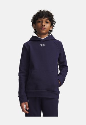 Poika, jolla on kiharat hiukset, pukeutunut tummansiniseen Under Armour -huppariin ja siihen sopiviin housuihin, seisoo valkoista taustaa vasten käsi taskussa.