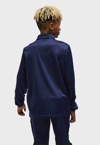 Person med blonda dreadlocks och rakade hårmönster bär en marinblå jacka med långa ärmar och matchande byxor, vänd bort från kameran.