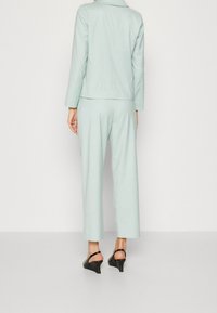 Vaaleanvihreä, räätälöity puku, jossa istuva bleiseri ja leveälahkeiset housut. Sileä pinta ja hillitty kiilto. Mustat korolliset kengät täydentävät asun.