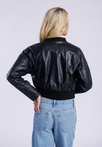 Černá kožená bomber bunda s žebrovanými manžetami a pasem, s malým embosovaným logem na zadní straně, kombinována se světlými modrými denimovými džíny.