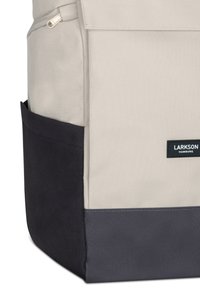 Sac à dos en tissu beige avec une section inférieure bleu foncé, comprenant une poche latérale et un patch logo. Fermeture zippée en haut.