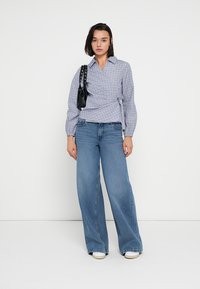 Μπλούζα τύπου wrap με καρό μοτίβο σε ανοιχτό μπλε, συνδυασμένη με φαρδιά μπλε τζιν παντελόνια. Μια μικρή μαύρη τσάντα με ροκ λουράκι αναδεικνύει την εμφάνιση.