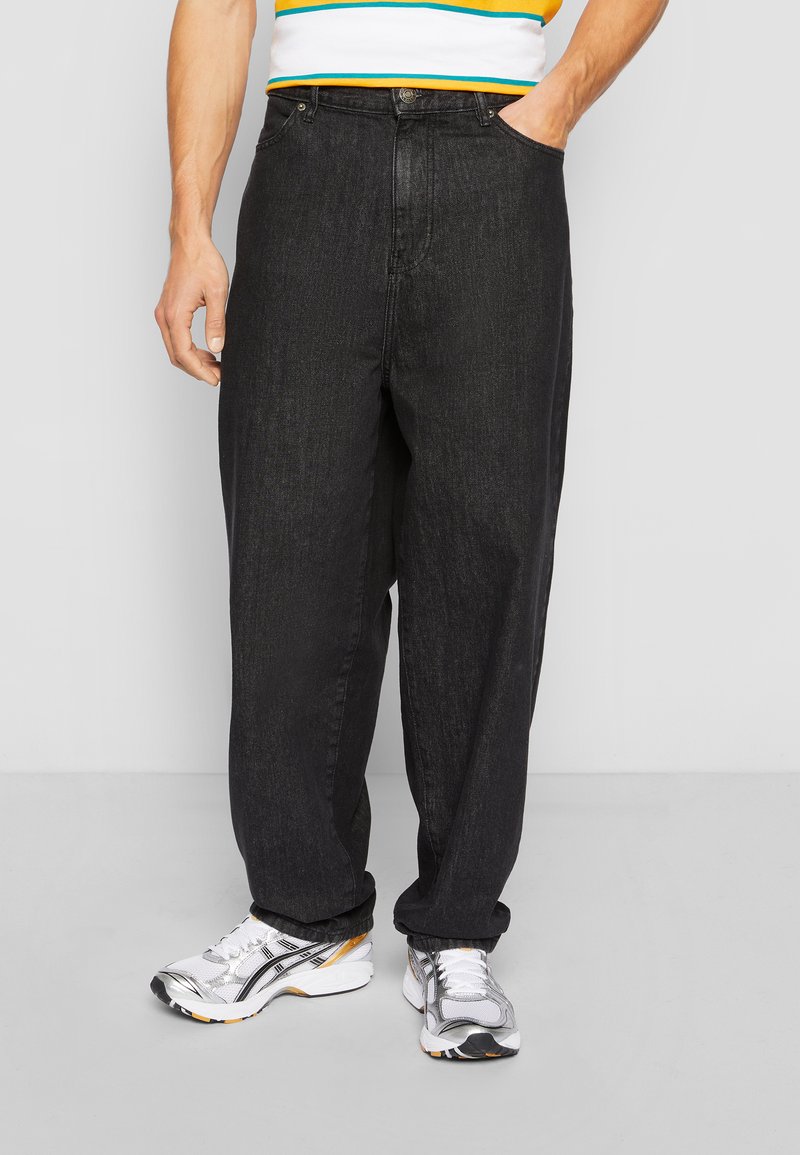Schwarze Denim-Jeans mit entspannter Passform, ausgestattet mit seitlichen Taschen und einem geraden Bein-Design. Der Stoff hat eine strukturierte Oberfläche, kombiniert mit silbernen Turnschuhen.