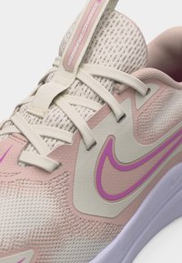 Κοντινό πλάνο ενός μπεζ και ροζ αθλητικού παπουτσιού Nike με διάτρητο επάνω μέρος, λευκά κορδόνια και μοβ λογότυπο Swoosh σε λευκό φόντο.