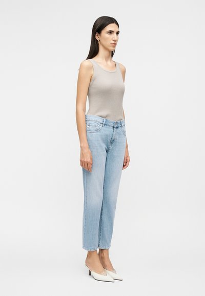 Γυναίκα που στέκεται με αμάνικο ανοιχτό γκρι τοπ, ανοιχτό μπλε ψηλόμεσα τζιν και λευκά μυτερά τακούνια σε απλό φόντο.