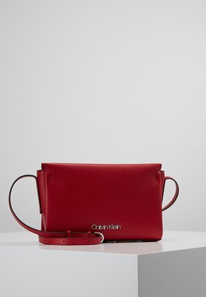 Rote Leder-Umhängetasche von Calvin Klein mit verstellbarem Riemen, minimalistischem Design und silbernem Logo auf der Vorderseite, präsentiert auf weißer Oberfläche.