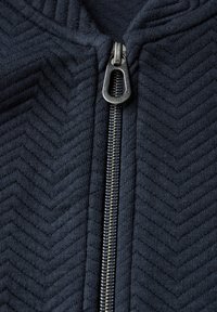 Giacca a zip navy con pattern a spina di pesce, dotata di zip metallica e texture del tessuto strutturata. Inquadratura ravvicinata dell'area della zip.