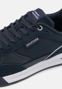Sneakers blu navy realizzati in tessuto e scamosciato, con suola bianca, dettagli testurizzati e un'etichetta "Hilfiger" sul lato.