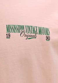 Grüner und schwarzer Text auf rosa Stoff lautet „MISSISSIPPI VINTAGE MOTORS Originals 1990“ in vintage-inspirierter Schriftart.