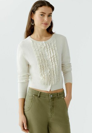 Light beige cardigan with ruffled front detail, featuring round neckline and buttons. Paired with olive green high-waisted pants.