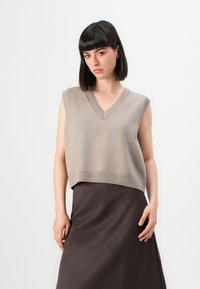 Donna con capelli scuri e lisci che indossa un gilet lavorato a maglia color beige senza maniche e una gonna marrone scuro su uno sfondo bianco semplice.
