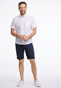 Camicia a maniche corte, rosa chiaro, con bottone e tasca sul petto, abbinata a pantaloni corti blu navy con risvolti; indossata con sneaker bianche.