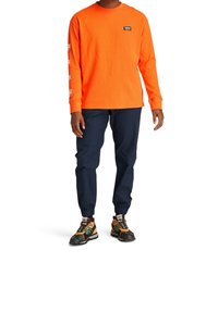 Orange långärmad skjorta med logotyp, kombinerad med marinblå joggerbyxor och flerfärgade, strukturerade skor med snörning och stötdämpande sulor.