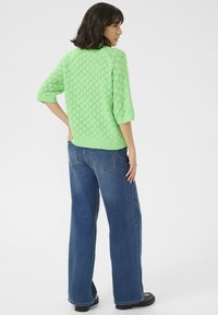Lichtgroene gebreide trui met een gestructureerd ruitpatroon, korte mouwen, gecombineerd met blauwe wijde jeans en zwarte schoenen.