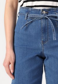 Jeans de ganga azul claro com cintura alta, apresentando um detalhe de laço na frente e um clássico design de cinco bolsos. Textura suave com costura discreta.