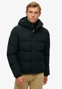 Kurtka puchowa w ciemnozielonym kolorze z kapturem, zapięciem na zatrzaski, pikowaną teksturą i bocznymi kieszeniami. Noszona na brązowej koszuli.