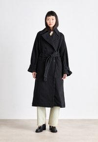 Manteau noir en trench avec un design croisé, grand col et ceinture nouée. Présente des poignets larges et une fente sur le côté, associé à un pantalon vert clair.