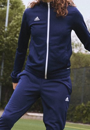 Persoon draagt een donkerblauw Adidas trainingspak met witte rits en logo's, staand buiten nabij een sportveld met groene achtergrond.