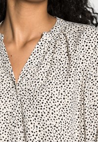 Helle Bluse mit einem gepunkteten Muster aus schwarzen Punkten, mit einem V-Ausschnitt und dezenten Raffungen an den Schultern. Sanfte Textur.