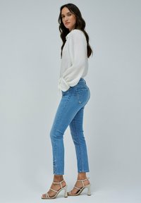 Blusa blanca de manga larga con puños atados, combinada con jeans ajustados de mezclilla azul claro con detalles de botones en el dobladillo y tacones altos beige con tiras.