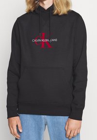 Felpa nera con cappuccio e tasca frontale a marsupio, caratterizzata da un prominente logo ricamato rosso "CK" e testo "CALVIN KLEIN JEANS" in bianco.