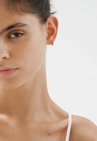 Pendiente de aro plateado exhibido en la oreja derecha de una persona. La piel parece suave con un tono natural; el cabello es oscuro y está recogido. Iluminación suave.