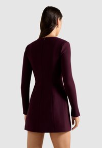 Langarm, getailleerde jurk van diep bordeauxkleurige stof. Heeft een ritssluiting aan de achterkant en subtiele naden, met een korte zoom.