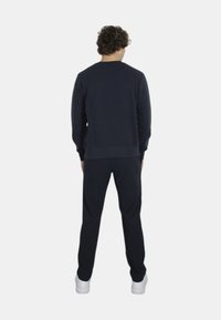 Maglione blu navy con scollatura rotonda e polsini a coste, abbinato a pantaloni della tuta coordinati. Modello visto da dietro, indossando sneakers bianche.