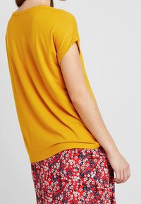 Top corto giallo senape a maniche corte con una vestibilità rilassata, abbinato a una gonna rossa con motivo floreale caratterizzata da piccoli fiori blu e bianchi.