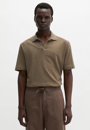 Mann trägt ein braunes kurzärmeliges Poloshirt und passende Hosen mit Kordelzug, steht vor einem schlichten weißen Hintergrund.