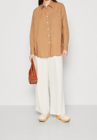 Camicia oversize marrone chiaro con bottoni bianchi, abbinata a pantaloni larghi bianchi e zoccoli di legno gialli. Porta una borsa in pelle marrone.