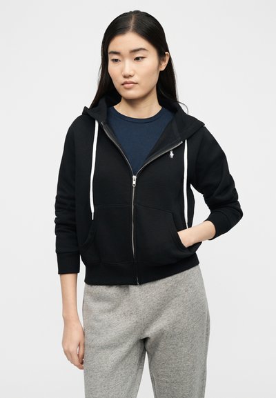 Νεαρή γυναίκα που φοράει μαύρο φούτερ με φερμουάρ πάνω από μπλε πουκάμισο και γκρι φόρμα, στέκεται με το ένα χέρι στην τσέπη του φούτερ, κοιτάζοντας προς τα κάτω.