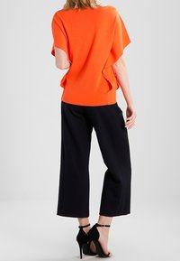 Pomarańczowa bluzka z krótkim rękawem i bocznymi rozcięciami, w połączeniu z czarnymi szerokimi spodniami i czarnymi sandałami na wysokim obcasie. Prosty fason, gładka faktura.