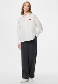 Camisa blanca de botones con un estampado de labios rojos, combinada con pantalones de pierna ancha gris oscuro y zapatillas blancas. Tejido ligero, corte relajado.
