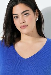 Maglione a V, blu vibrante, in maglia con una fine trama. La modella indossa orecchini a cerchio in argento e un trucco naturale. Lo sfondo è neutro.