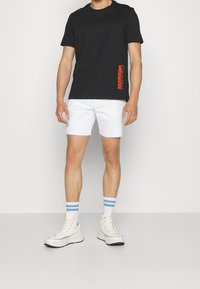 Svart bomullströja med röd logga, vita denimshorts med fransar, och högstövlar i vitt med randiga blå strumpor.