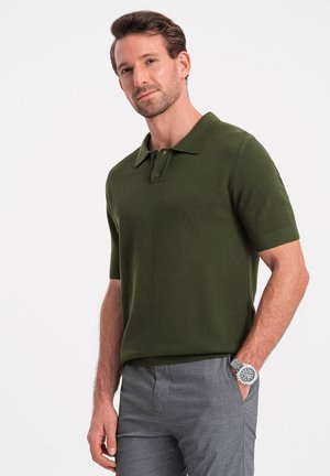 Polo shirt lavorato a maglia verde scuro con colletto classico, maniche corte e un motivo testurizzato, abbinato a pantaloni grigi.
