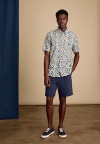 Korte mouw overhemd met een bloemenpatroon in geel, blauw en groen. Gecombineerd met marineblauwe shorts en marineblauwe schoenen met witte veters.