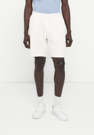 Uomo che indossa pantaloncini bianchi con coulisse, calzini bianchi e scarpe da ginnastica bianche, in piedi davanti a uno sfondo bianco semplice.