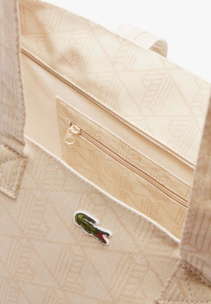 Beige Stofftasche mit einem Reißverschlussfach innen und einem gestickten grünen Krokodil-Logo auf der Vorderseite.