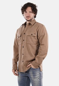Camicia in flanella marrone con finitura testurizzata, due tasche frontali, chiusura a bottoni e collo classico, abbinata a jeans in denim sfilacciati.