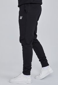 Zwarte joggings met een zachte stof, voorzien van zijzakken, geribbelde boorden en een wit logo op de linker bovenbeen. Gecombineerd met witte sneakers.