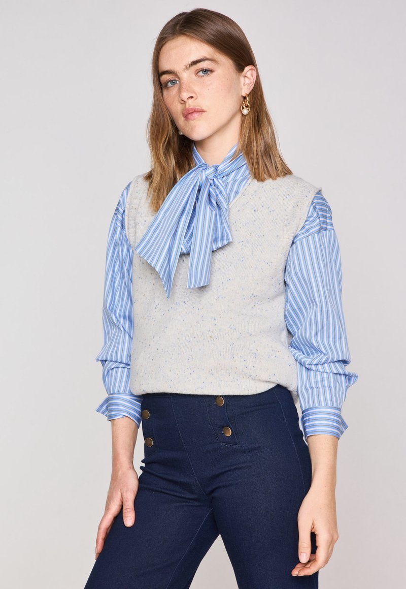 Gilet a maglia grigia a pois sopra una camicia a righe azzurra e bianca con cravatta a farfalla. Pantaloni a vita alta blu scuro con accenti di bottoni in ottone.