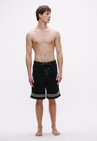 Μαύρα μαγιό σορτς με κίτρινα ράμματα, ελαστική μέση με κορδόνι και σχεδιασμό μέχρι το γόνατο. Το μοντέλο στέκεται ξυπόλητο.