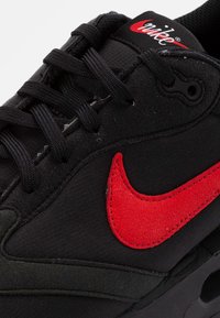 Zwarte sneaker gemaakt van mesh en suède, met een opvallende rode Nike swoosh, zwarte veters en een getextureerde tussenzool.