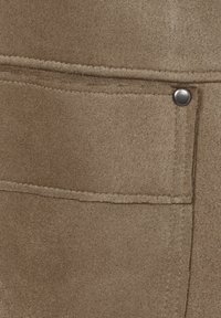 Beiger Stoff mit weicher Textur und einer aufgesetzten Tasche, die durch einen Metallschnappverschluss gesichert ist. Sichtbare Nähte fügen entlang der Kanten Details hinzu.