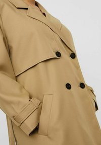 Beige Trenchcoat mit doppelreihigem Design, schwarzen Knöpfen, breiten Revers und seitlichen Taschen. Das Material wirkt glatt und robust.