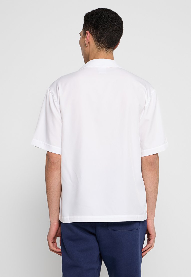 Hombre con una camisa blanca de manga corta y pantalones azul oscuro, de pie de espaldas contra un fondo gris claro liso.