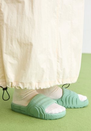 Ljusa myntgröna sandaler med en strukturerad design och breda remmar, tillsammans med vita ankelsockar, under ett ljust beige plagg.